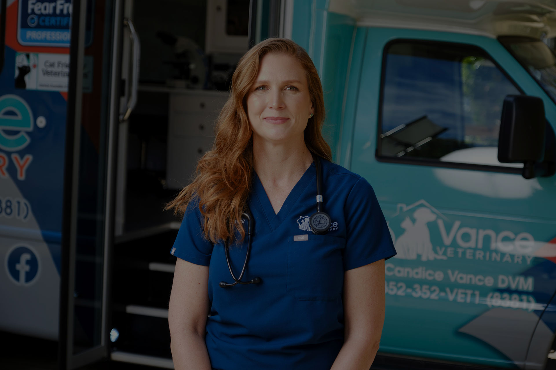 A veterinarian in blue scrubs with a stethoscope around her neck stands in front of a veterinary mobile clinic with "Vance Veterinary" written on the vehicle.