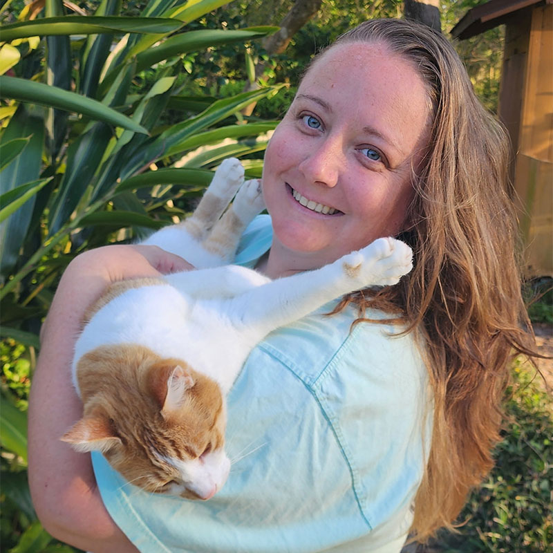 A smiling veterinarian with long brown hair holds a relaxed orange and white cat outdoors, standing near green plants and a wooden structure in the background.