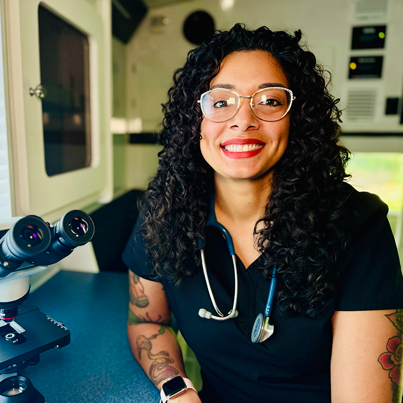 A smiling veterinarian with curly hair, glasses, and a stethoscope around her neck sits beside a microscope in a medical setting. She is wearing black scrubs and has visible tattoos on her arms.
