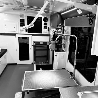 Black and white image of a clean, modern veterinary surgery room inside a mobile clinic, featuring an exam table, surgical lights, medical equipment, and cabinets—a perfect workspace for any veterinarian—with a window on the right side.