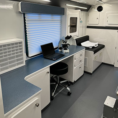 A clean, modern mobile veterinary laboratory with a blue countertop, microscope, laptop, storage drawers, and a black rolling chair. There is a padded bench along the wall and a window with a closed blind—ideal for any vet on the go.