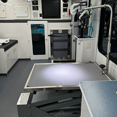 A clean, modern veterinarian exam room with an adjustable exam table in the center, medical equipment, monitors, and cabinets lining the walls. The vet’s workspace is well-lit and organized.