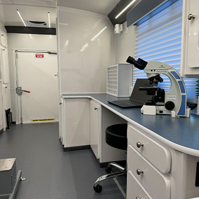 A modern, clean veterinary laboratory workspace with white cabinets, blue countertops, a microscope, a laptop, and a stool. The room has a window with blinds and a door at the back with warning signs for vet staff.