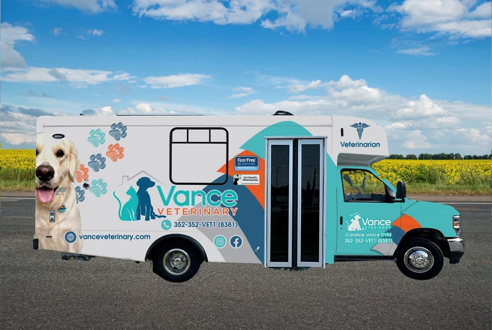 A mobile veterinarian clinic van parked on a rural road, decorated with paw prints, a dog’s face, and the Vance Veterinary branding on the side, stands out against green fields and a blue sky.